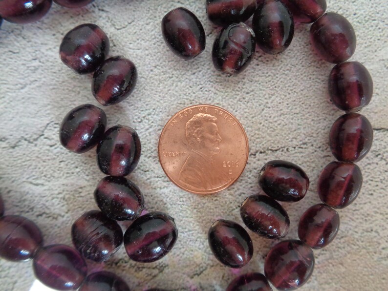 May include: A strand of dark purple glass beads, some of which are oval shaped and some are round. The beads are arranged in a circle around a penny.