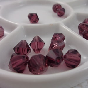 May include: A white plastic tray containing a selection of small, faceted, purple glass beads. The beads are arranged in a circular pattern.