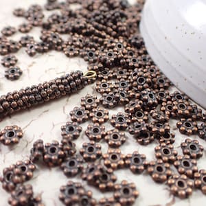 May include: A large quantity of small, copper-colored metal beads with a flower-shaped design. The beads have a hole in the center for stringing.