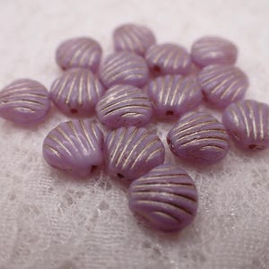 May include: A collection of lavender-colored shell-shaped beads with gold-colored stripes. These beads are small, with a hole for stringing. The beads are scattered on a white, textured surface. Ideal for jewelry making.