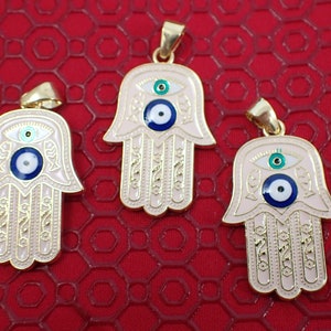 Op de afbeelding: Drie gouden hangers met een roze emaille afwerking. Elke hanger heeft een hamsa hand ontwerp met een blauw boze oog in het midden. Het boze oog heeft een witte kern en een groene buitenring.