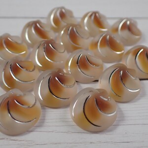 May include: A collection of 16 swirl-shaped buttons in a light brown and white color scheme. The buttons have a raised, textured design and a silver-colored outline.