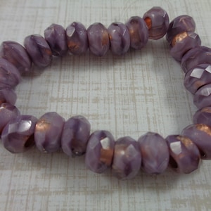 May include: A strand of faceted, lavender-colored beads with copper-colored accents. The beads are oval-shaped and have a polished, reflective surface. The beads are strung together, forming a bracelet or necklace.