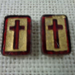 May include: Two small, square, red glass tiles with gold crosses inside. The crosses are raised and have a slightly textured surface.