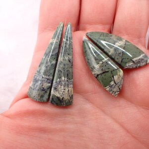 May include: Four polished, triangular-shaped cabochons made of green stone with dark grey and white veining. The stones feature a smooth, glossy surface with distinct natural patterns of intersecting lines and varied shades of sage and olive green.