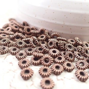 May include: A pile of copper-colored metal beads with a decorative, ridged, flower-like design. The beads are small and round with a hole in the center.