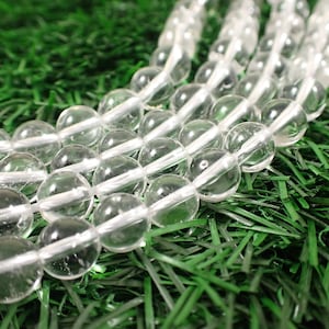 May include: A strand of clear quartz beads, each bead is round and smooth. The beads are arranged in a line on a green surface.