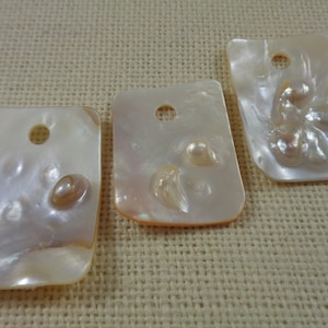 May include: Three rectangular mother-of-pearl pendants with a hole at the top. Each pendant features a unique arrangement of natural pearl formations. The pendants have an iridescent sheen with hints of pink and cream.