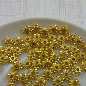 May include: A pile of gold-colored metal beads with a decorative design. The beads have a round hole in the center and are arranged in a random pattern.