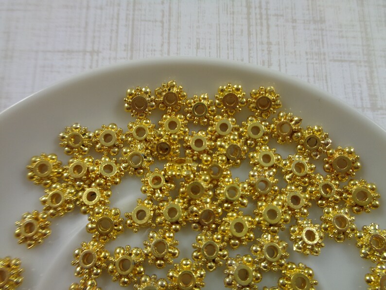 May include: A pile of gold-colored metal beads with a decorative design. The beads have a round hole in the center and are arranged in a random pattern.