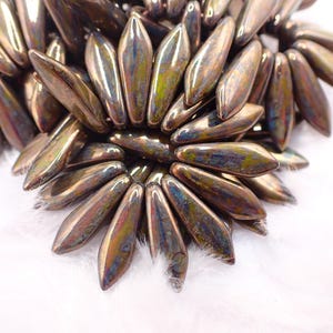 May include: A collection of elongated, petal-shaped beads with a metallic, iridescent finish. The beads feature a blend of bronze, copper, and subtle green and blue hues, arranged in a dense, layered cluster that creates a textured, radial pattern.