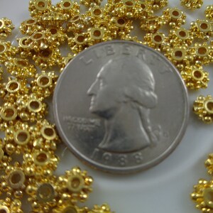 May include: A close-up of a pile of gold-colored metal beads with a starburst design. The beads are arranged around a US quarter, which is used to show the size of the beads.
