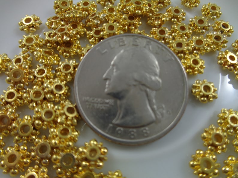 May include: A close-up of a pile of gold-colored metal beads with a starburst design. The beads are arranged around a US quarter, which is used to show the size of the beads.