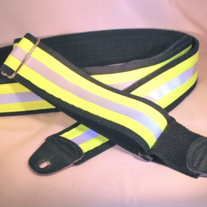 May include: A black guitar strap with a neon yellow and reflective silver stripe design. The strap is padded for comfort and has black leather ends with a hole for attaching to a guitar.