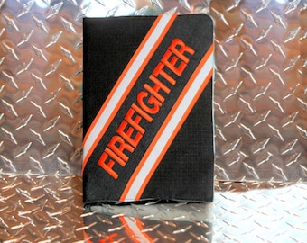 Op de afbeelding: Zwarte notitieboekomslag met oranje en witte diagonale strepen en het woord "FIREFIGHTER" geborduurd in oranje.