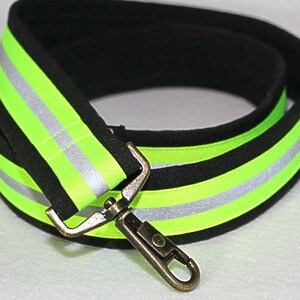 May include: A black and green reflective strap with a silver clip. The strap is made of a soft, padded material and has two reflective stripes.