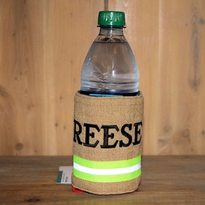 May include: A tan fabric can cooler with a reflective yellow stripe and the name "REESE" embroidered in black. A clear plastic water bottle with a green lid is inside the cooler.