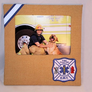 May include: A framed photo of a firefighter with a dog in front of a fire truck. The frame is made of brown fabric and has a blue and white ribbon on the top left corner. The photo shows a firefighter in a yellow helmet and a dog with its tongue sticking out. The frame also has a blue and white patch with the words "Fire Fighter" and "E.M.T." on it.