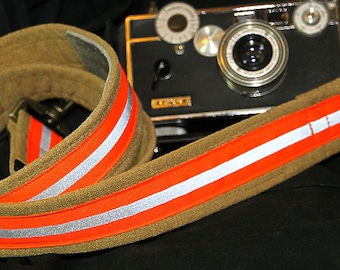 May include: A brown camera strap with two orange and white reflective stripes. The strap is made of a thick, durable material and is designed to be comfortable to wear.