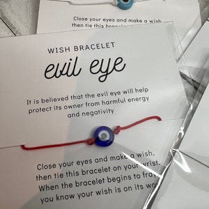 May include: Red string bracelet with a blue evil eye charm. The bracelet is tied around a white card with the text "WISH BRACELET evil eye" and instructions on how to make a wish.