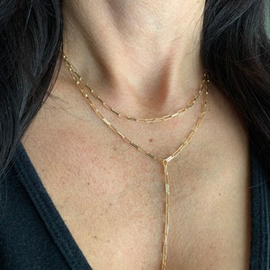 May include: A gold-coloured necklace with two strands. The top strand is a simple chain, and the second has a vertical drop. The necklace is worn against a black top.