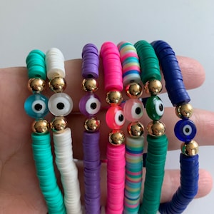 May include: Set of eight colourful bracelets with evil eye charms. The bracelets are made of small, round beads in various colours, including turquoise, white, purple, pink, and green. Each bracelet has a gold bead and a blue evil eye charm.