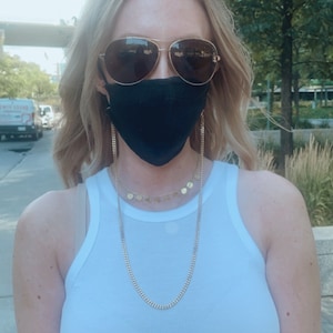 May include: A woman wearing a white tank top and black face mask with a gold chain attached. The chain has small gold circles hanging from it.