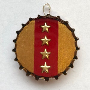 Könnte beinhalten: Goldener Flaschenverschluss mit einem roten Streifen und vier goldenen Sternen. Der Flaschenverschluss hat eine silberne Schlaufe zum Aufhängen.