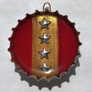 Könnte beinhalten: Ein roter Flaschenverschluss mit einem goldenen Streifen und vier silbernen Sternen. Der Flaschenverschluss hat eine Kupferschlaufe zum Aufhängen.