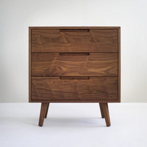 May include: A three-drawer wooden dresser with a warm brown finish. The drawers have recessed handles and the dresser stands on four angled legs. The wood grain is visible, adding texture to the piece. The design is modern and minimalist.
