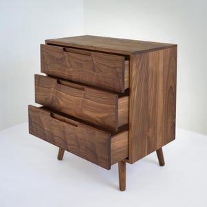 May include: A three-drawer wooden dresser with a mid-century modern design. The dresser is made of dark brown wood with visible grain and features three drawers with integrated handles. The drawers are partially open, revealing the interior. The dresser stands on four tapered legs.