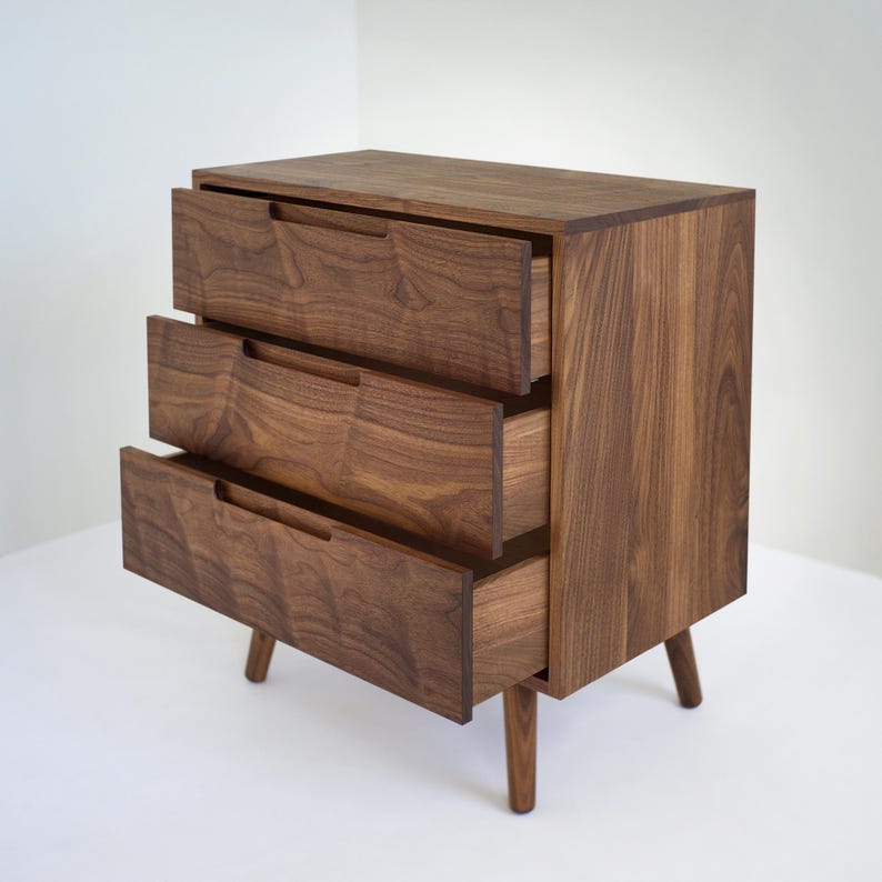 May include: A three-drawer wooden dresser with a mid-century modern design. The dresser is made of dark brown wood with visible grain and features three drawers with integrated handles. The drawers are partially open, revealing the interior. The dresser stands on four tapered legs.