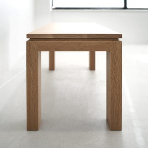 May include: A wooden bench with a natural finish and a rectangular top. The bench has four legs and a simple design.