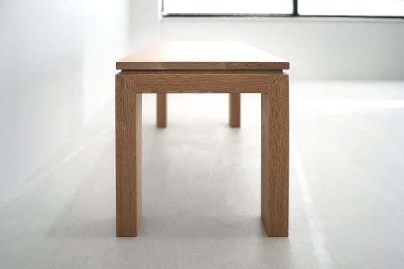 May include: A wooden bench with a natural finish and a rectangular top. The bench has four legs and a simple design.