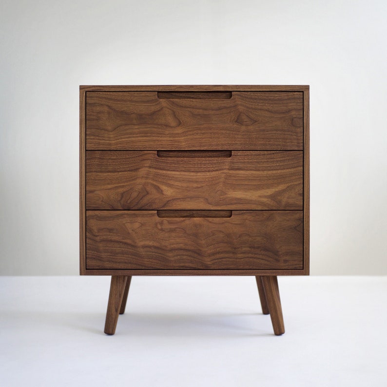 May include: A three-drawer wooden dresser with a warm brown finish. The drawers have recessed handles and the dresser stands on four angled legs. The wood grain is visible, adding texture to the piece. The design is modern and minimalist.