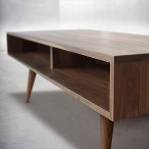 May include: A wooden coffee table with a rectangular top and two open shelves. The table has a light brown finish and is supported by four tapered legs.