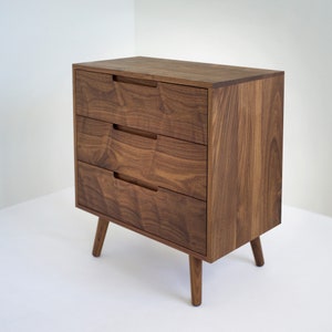 May include: A mid-century modern style wooden dresser with three drawers. The dresser is made of dark brown wood with visible grain and has recessed drawer pulls. It stands on four tapered legs.