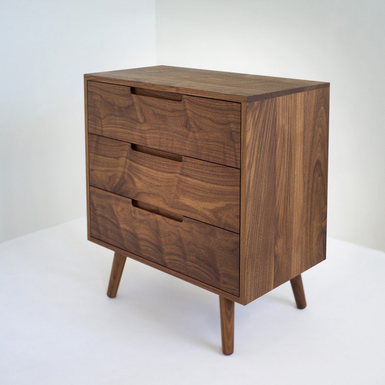 May include: A mid-century modern style wooden dresser with three drawers. The dresser is made of dark brown wood with visible grain and has recessed drawer pulls. It stands on four tapered legs.