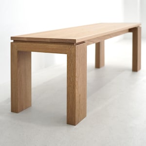 May include: A light brown wooden bench with four legs. The bench is made of solid wood and has a simple, modern design.