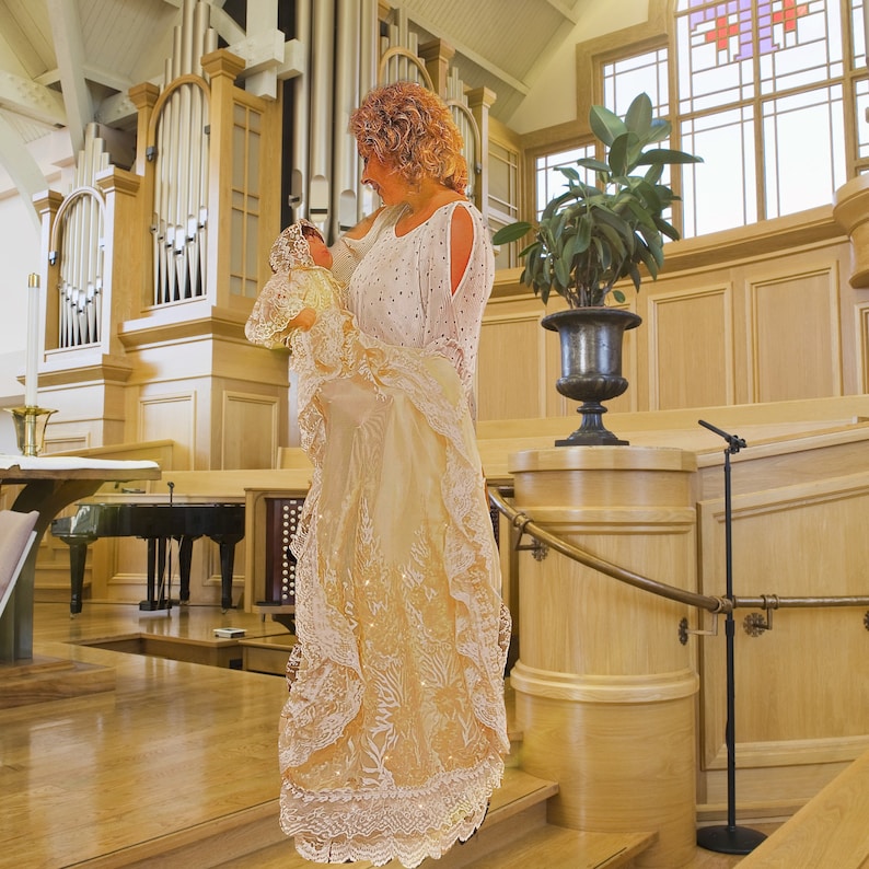 May include: A cream-coloured christening gown with lace trim and a matching bonnet. The gown features a delicate floral pattern and is held by a person in a white dress with cut-out sleeves. The background includes a church interior.