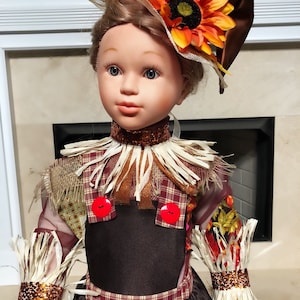 May include: A decorative doll dressed in a fall-themed outfit. The doll wears a brown dress with a plaid bodice, straw accents, and a hat adorned with a sunflower. The doll has a fair complexion and blue eyes.
