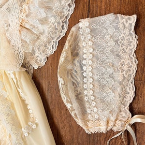 May include: A cream-coloured baby dress and bonnet set. The dress features delicate lace detailing on the sleeves and bodice. The bonnet is also adorned with lace and has ribbon ties. The set is made of a soft, sheer fabric.