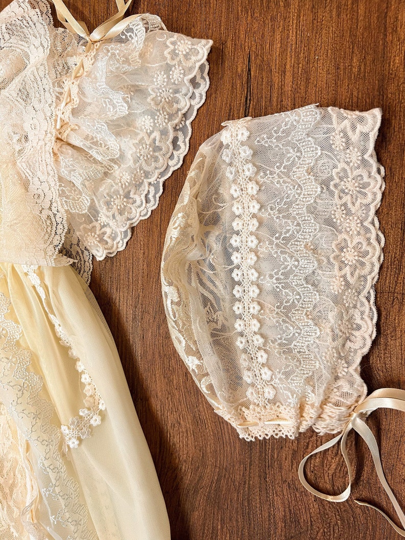 May include: A cream-coloured baby dress and bonnet set. The dress features delicate lace detailing on the sleeves and bodice. The bonnet is also adorned with lace and has ribbon ties. The set is made of a soft, sheer fabric.