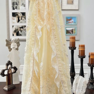 May include: A cream-coloured christening gown with lace trim and a matching bonnet. The gown features a floral design and is hanging on a hanger. The words "With Flower Belt" and "Royal Christening Gown" are written above the gown.