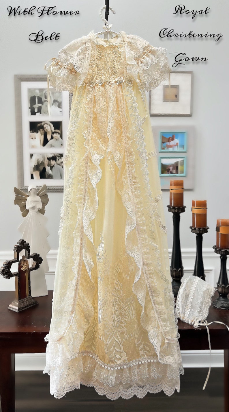 May include: A cream-coloured christening gown with lace trim and a matching bonnet. The gown features a floral design and is hanging on a hanger. The words "With Flower Belt" and "Royal Christening Gown" are written above the gown.