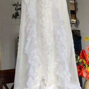 May include: A white christening gown and bonnet set. The gown features intricate lace detailing, ruffles, and a delicate design. The bonnet has a matching lace trim and ribbon ties. The set is displayed on a wooden surface.