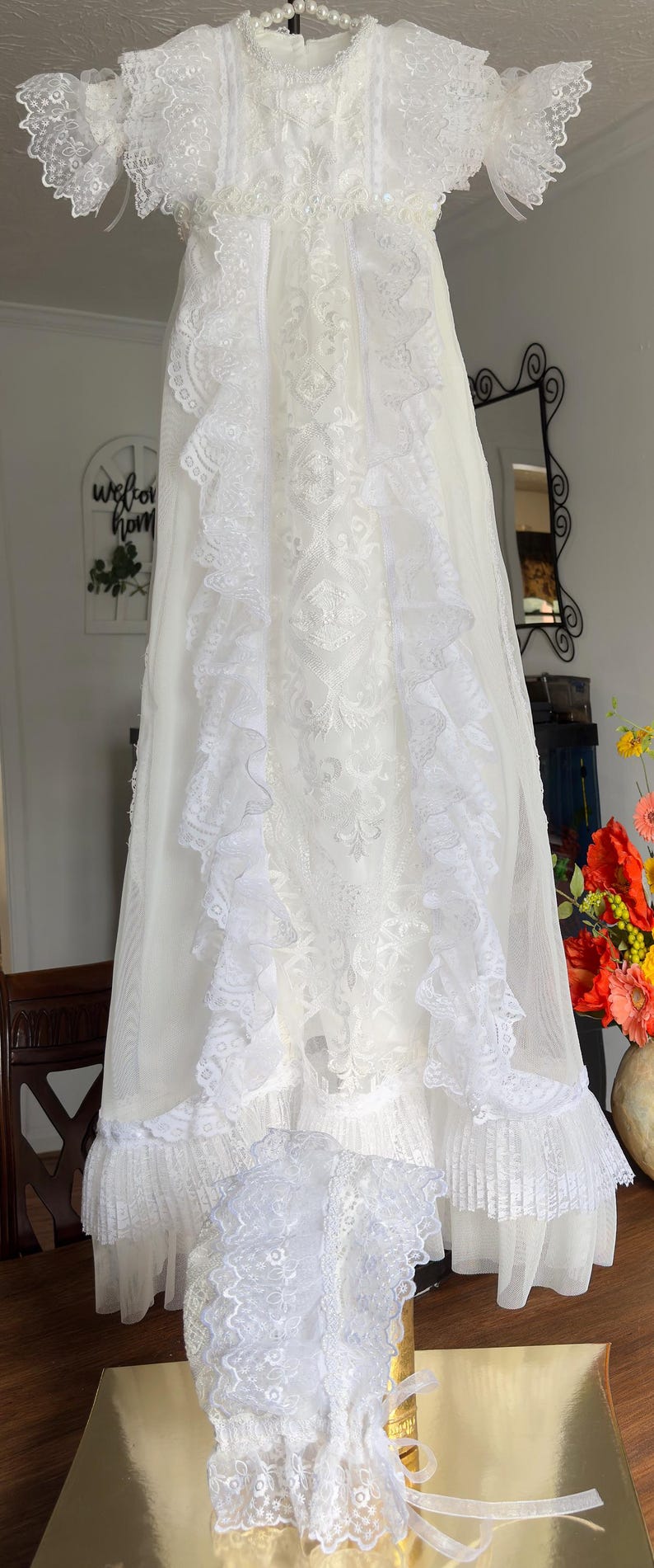 May include: A white christening gown and bonnet set. The gown features intricate lace detailing, ruffles, and a delicate design. The bonnet has a matching lace trim and ribbon ties. The set is displayed on a wooden surface.