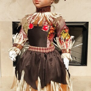 May include: A decorative doll dressed as a scarecrow, featuring a brown satin skirt, a black bodice, and a straw-like collar and cuffs. The doll wears a hat adorned with a large orange sunflower and a brown, pointed brim. The doll is wearing white gloves and white stockings.