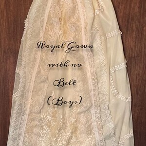 May include: Cream-coloured christening gown with lace detailing. The gown features a delicate floral pattern, lace trim, and a ruffled hem. The text on the gown reads "Royal Gown with no Belt (Boys)".