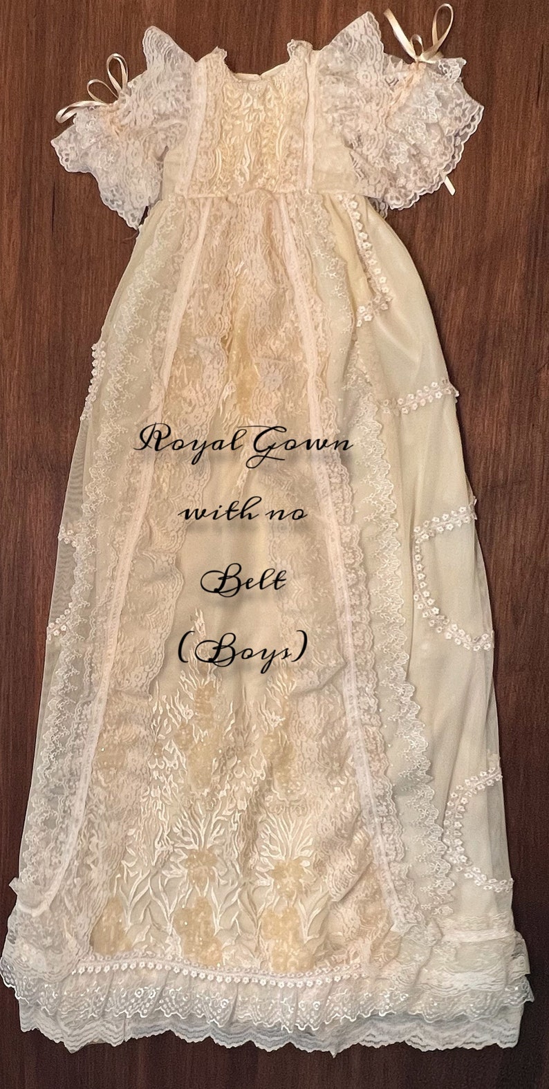 May include: Cream-coloured christening gown with lace detailing. The gown features a delicate floral pattern, lace trim, and a ruffled hem. The text on the gown reads "Royal Gown with no Belt (Boys)".